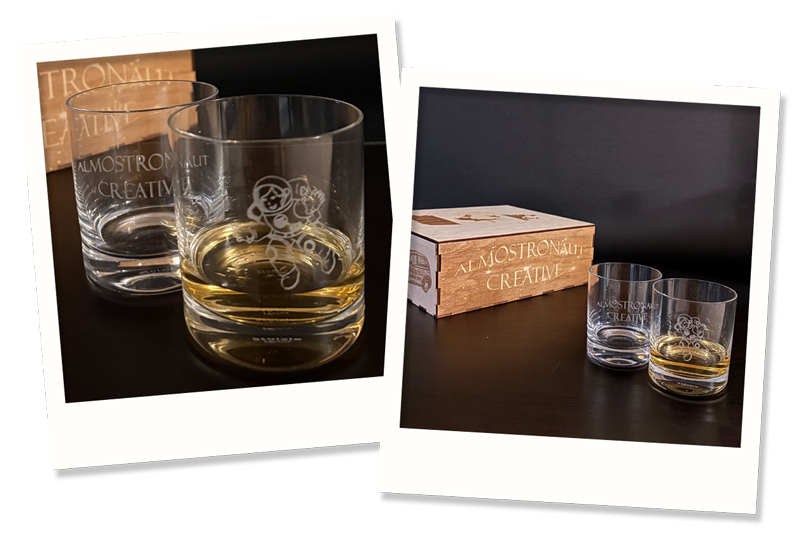 Two clear whisky glasses with etched designs, one filled with amber liquid, are placed next to a wooden box labeled "Almostronaut Creative" on a dark surface.