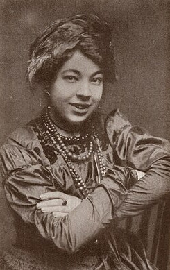 A sepia-toned portrait of a woman wearing a feathered hat, a satin dress, and multiple beaded necklaces, seated with her arms crossed and smiling slightly at the camera.