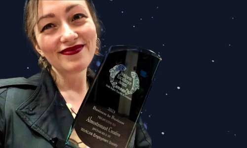 Marleen, a smiling woman with red lipstick and earrings, is holding the business excellence award plaque.